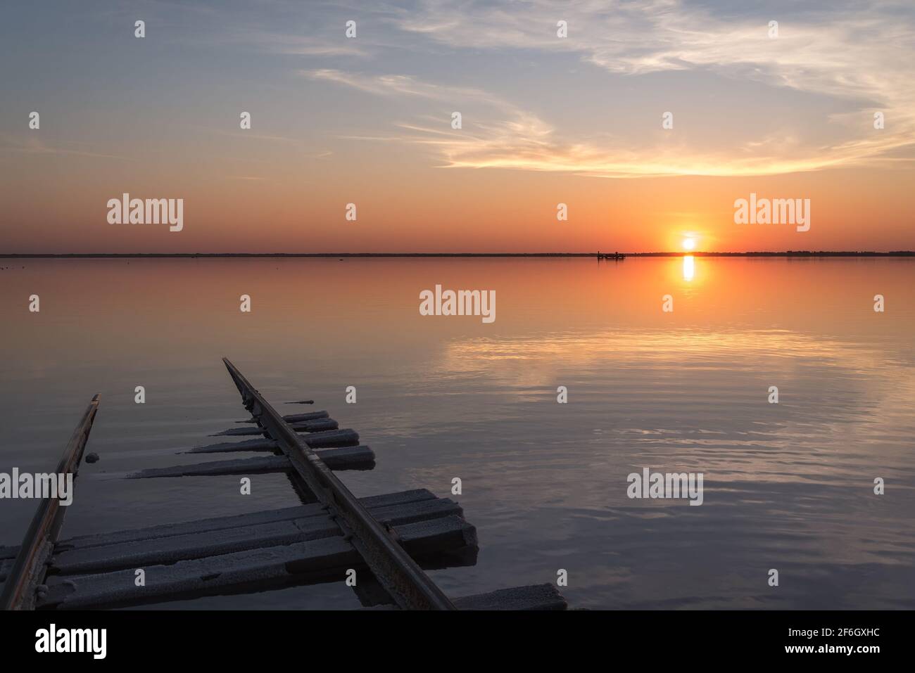 Erstaunlicher Sonnenuntergang über einem Salzsee mit wunderschönen Wolken, Sonne, Zug am Horizont, Reflexionen im Wasser und Schienen im Vordergrund. Lake Bursol, EIN Stockfoto