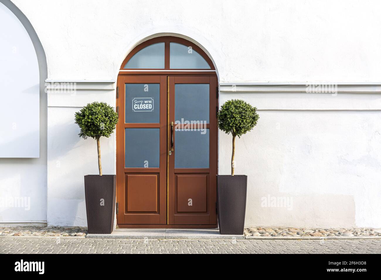 Geschlossene rote Holztür mit zwei dekorativen Bäumen in Töpfen an einer weißen Wand der Gebäudefassade. Restaurant, vorübergehend während Coronavirus pandemi geschlossen Stockfoto