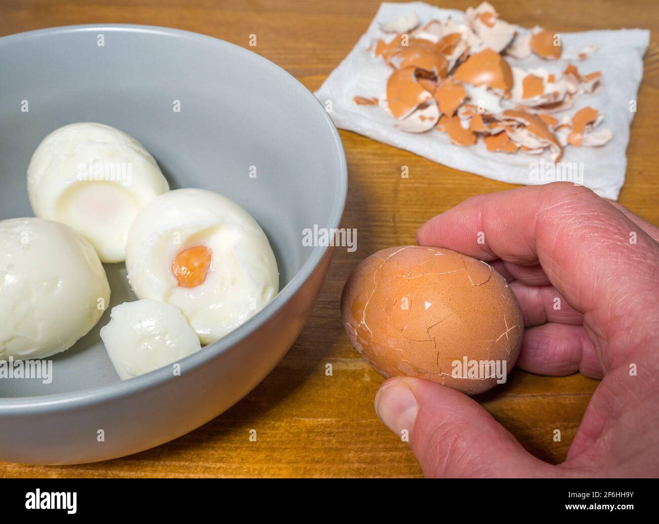 Nahaufnahme der Hand eines Mannes, die ein hart gekochtes Ei auf einer Kiefer-Arbeitsplatte / Theke knackt, neben einigen entfernten Schalen auf einer Serviette und einer Schüssel geschälte Eier. Stockfoto