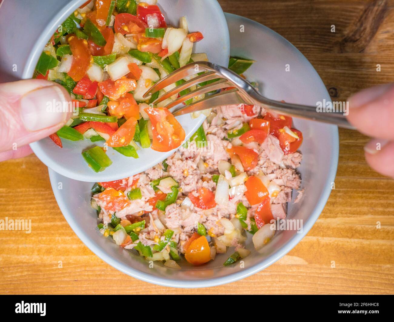Nahaufnahme eines POV-Schusses mit gehacktem Pfeffer, Zwiebeln und Tomaten, der mit einer Gabel aus einer kleinen Schüssel in eine größere Schüssel gehacktem Thunfisch gegossen wird. Stockfoto