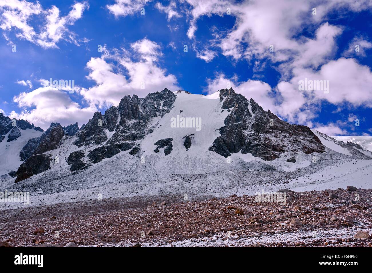 Felsiger Gipfel mit einem mächtigen Gletscher, der über dem schneebedeckten Berg thront Moräne im Hochland auf dem Hintergrund eines blauen Himmel mit Wolken Stockfoto