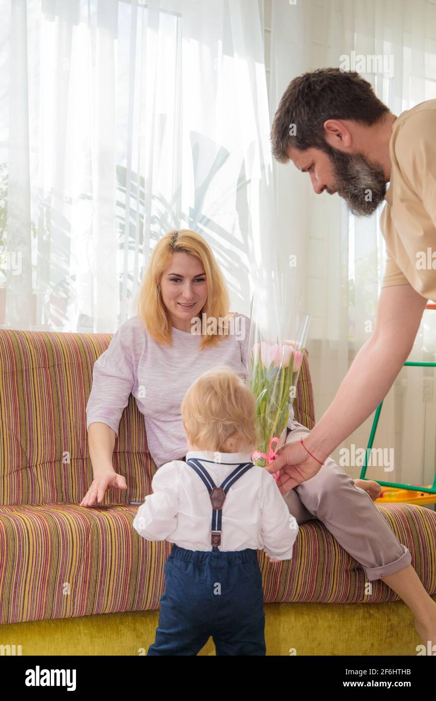 Papa mit Baby, Sohn gratuliert Mama zum Urlaub. Das Kind bringt seiner Mutter einen Blumenstrauß und ein Geschenk. Stockfoto