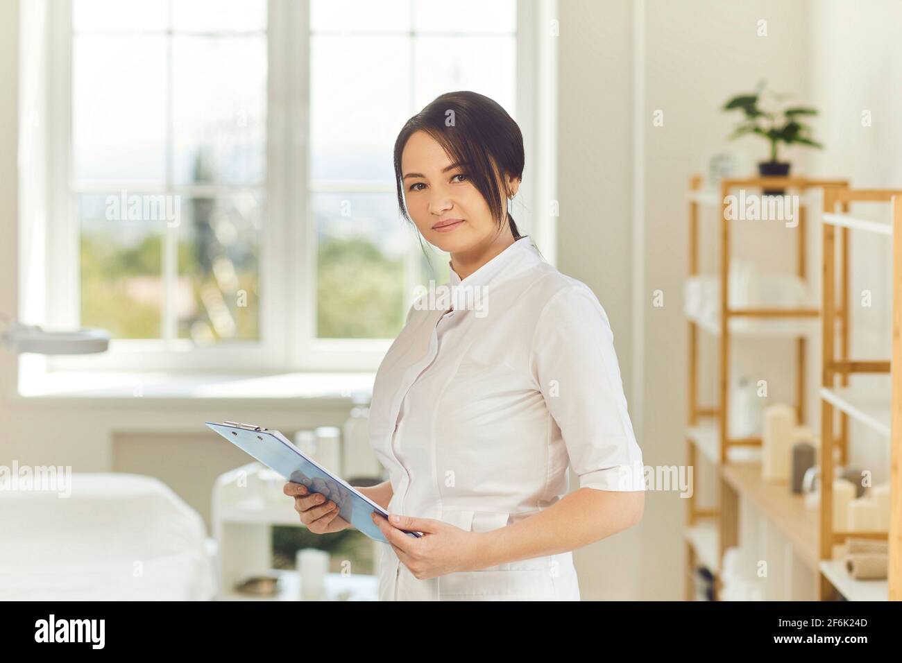 Lächelnde Frau Dermatologe in medizinischen Uniform stehend mit Dokumenten in Schönheitssalon Stockfoto