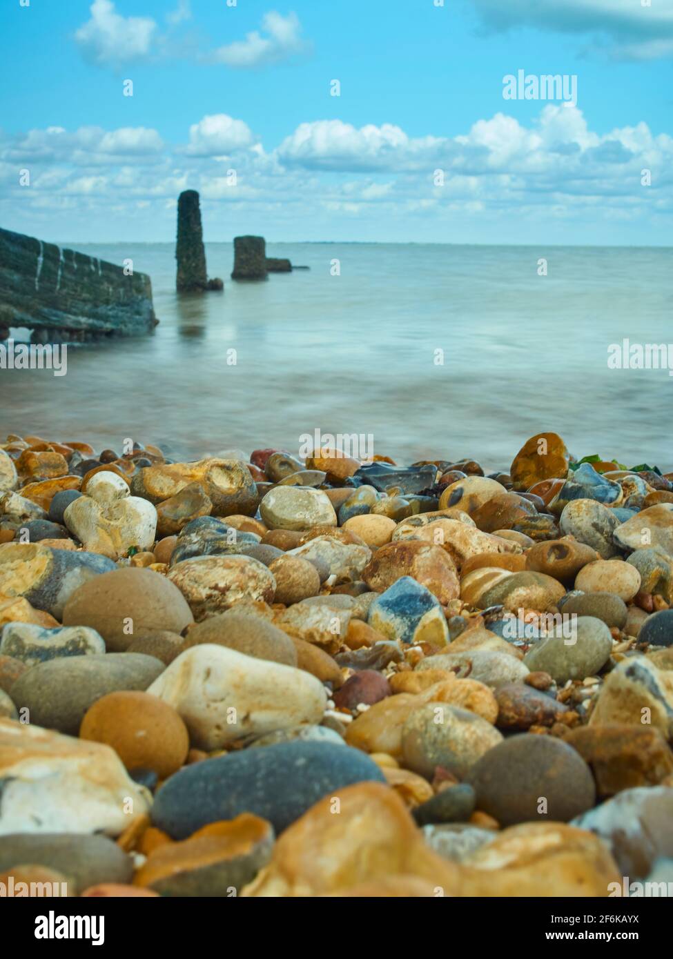 Ein Vordergrund aus Kieselsteinen, vor einem sich bewegende Meer mit einem verwelkenden Groyne in abnehmender Perspektive und einem Horizont mit hellem, blauem Sommerhimmel. Stockfoto