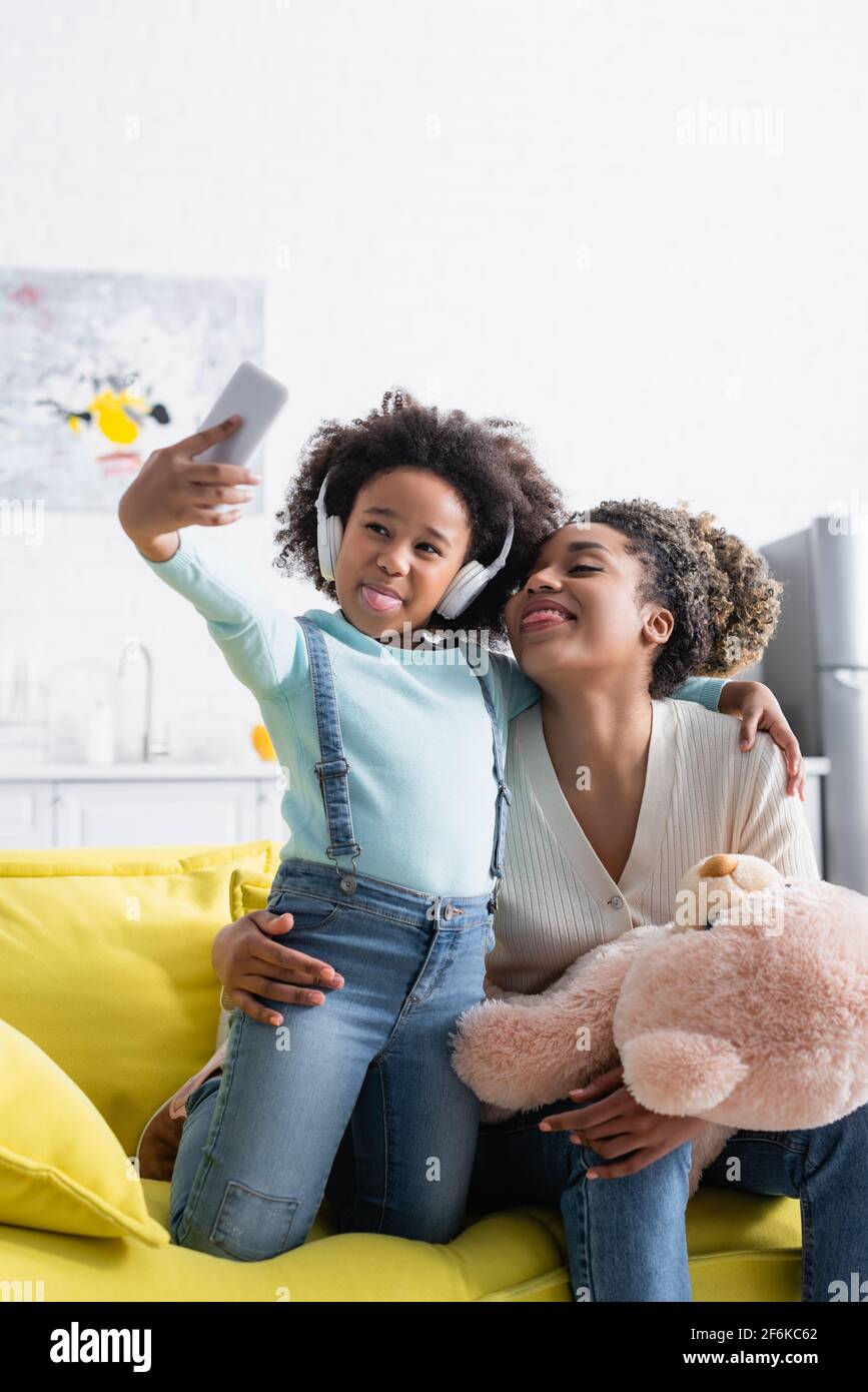 afroamerikanisches Mädchen mit Kopfhörern und Frau mit Teddybär Selfie machen und dabei Zungen herausstechen Stockfoto