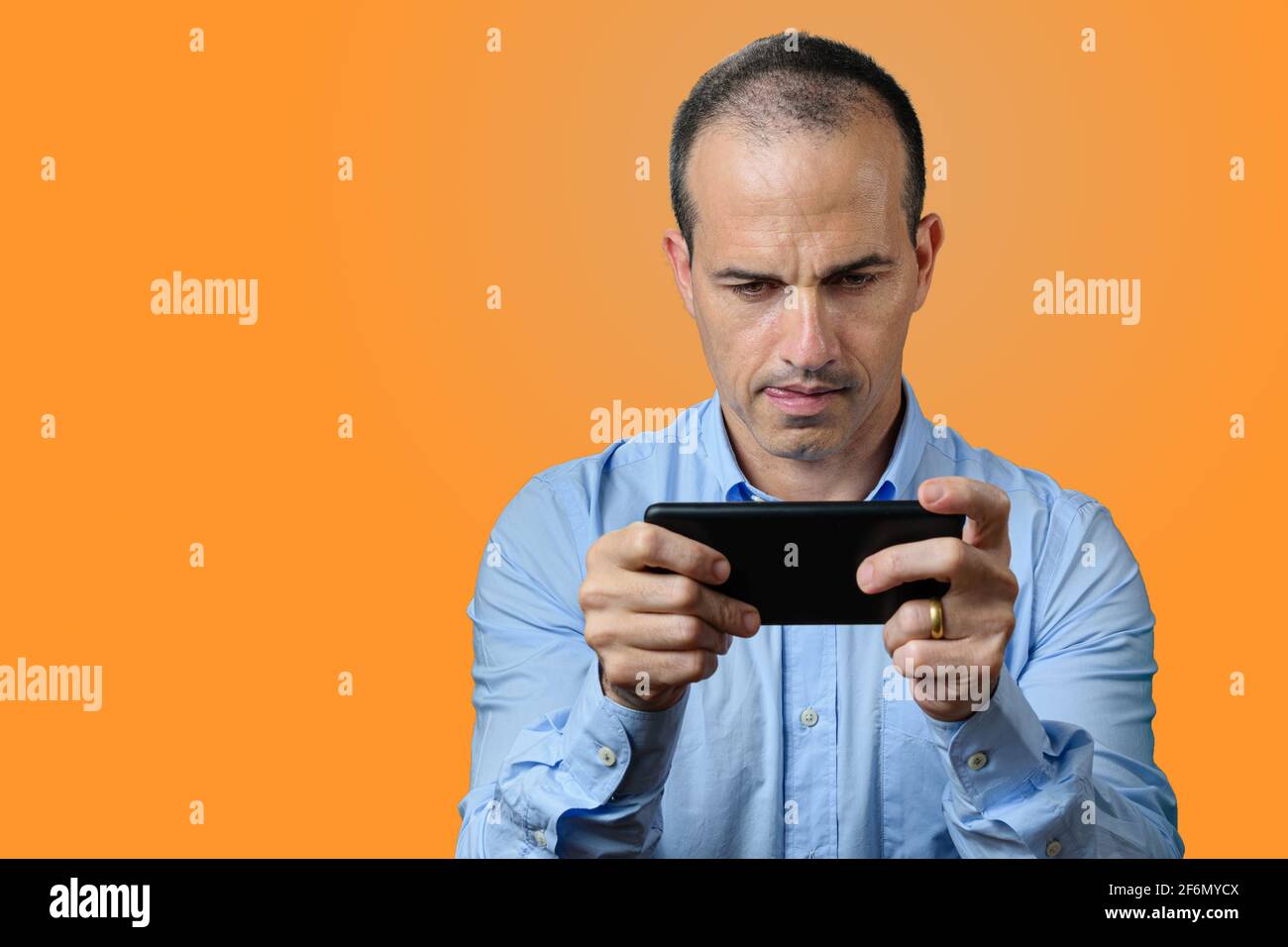 Reifer Mann in formeller Kleidung beißt seine Zunge und hält das Smartphone horizontal. Orangefarbener Hintergrund Stockfoto