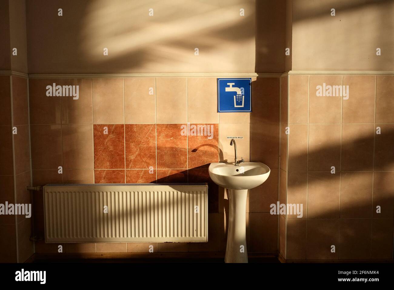 Weißes Waschbecken in der Nähe einer gefliesten Wand und blaues Zapfschild in einer Wartehalle des Bahnhofs, Pomitna, Ukraine. Kontrast zwischen Morgenlicht und Schatten Stockfoto