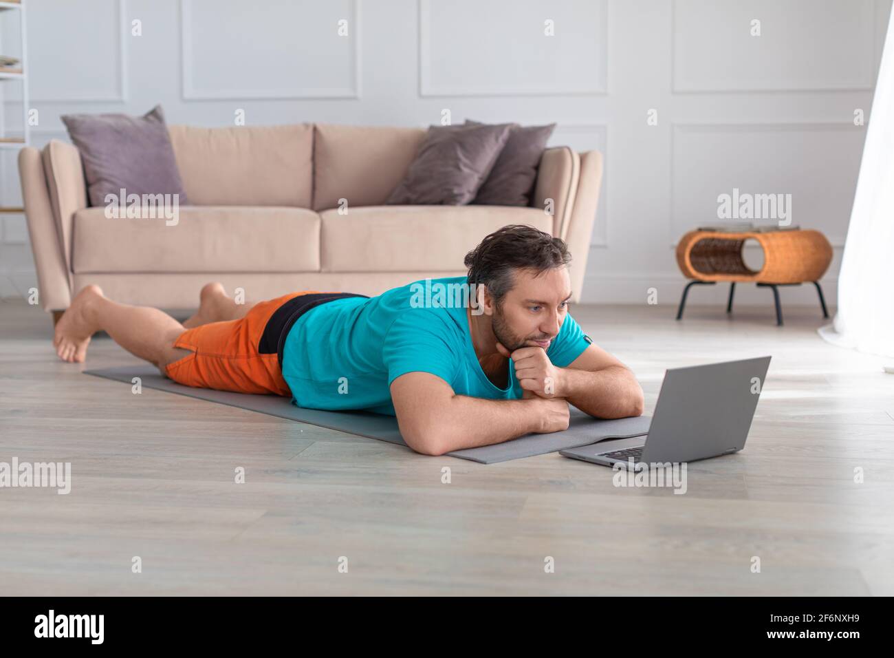 Ein Mann mittleren Alters schaut sich ein Yoga-Training Tutorial an. Er liegt zu Hause auf dem Boden vor einem Laptop-Monitor. Gesundes Lifestyle-Konzept. Stockfoto