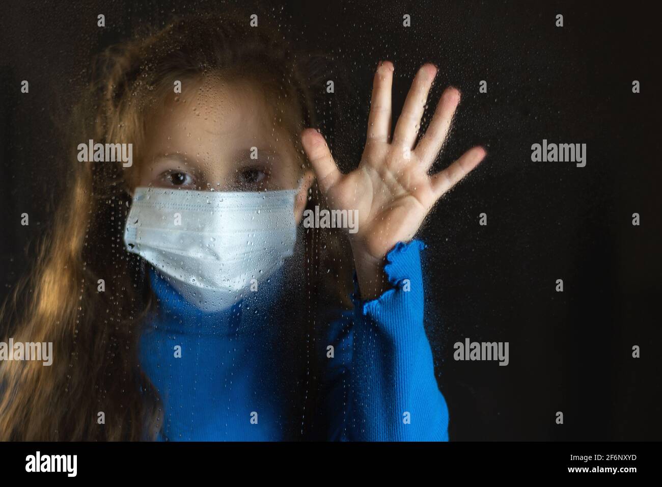 Baby Mädchen in medizinische Maske hinter nassem Glas.Tropfen auf Glas. Dunkler Hintergrund Stockfoto