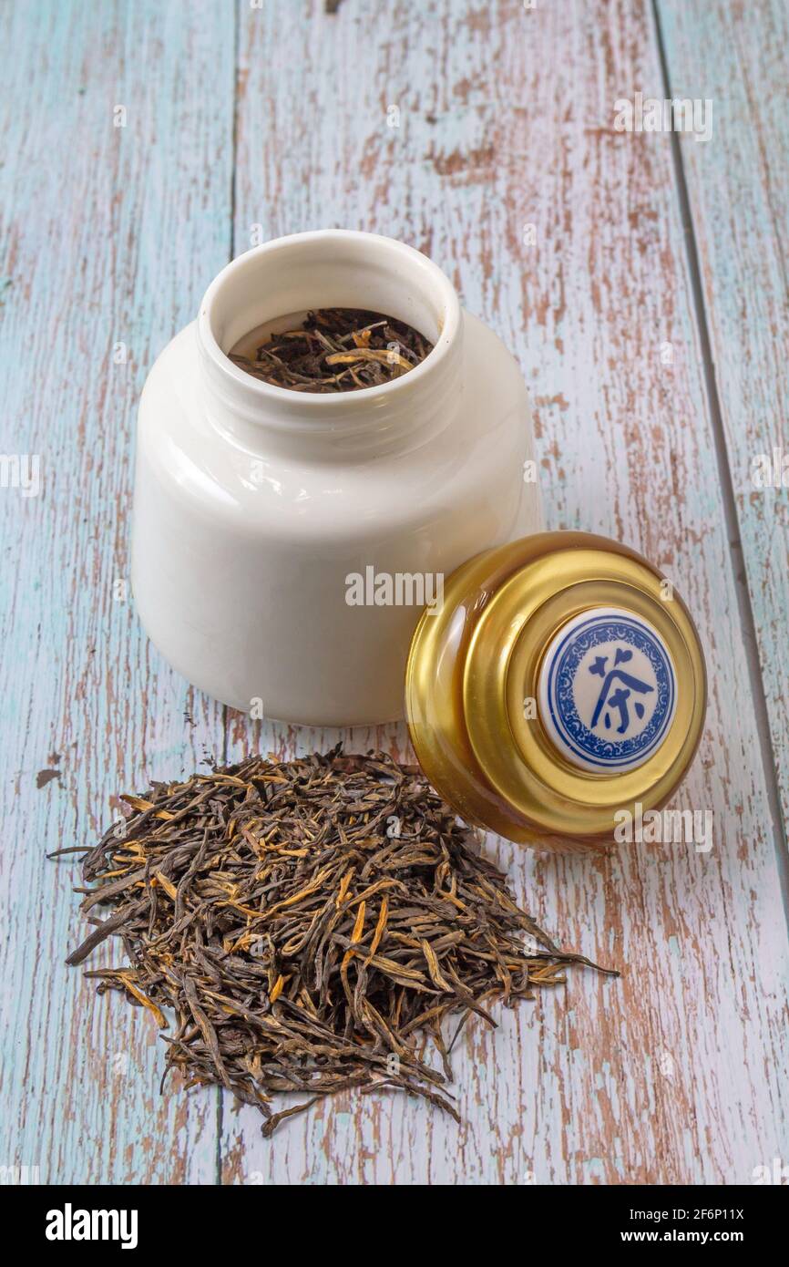 Teeblätter bereit für die Infusion und eine weiße Kanne, die Tee im Inneren enthält. Der Deckel, der auf dem Topf ruht, hat das Wort Tee auf Chinesisch geschrieben. Stockfoto