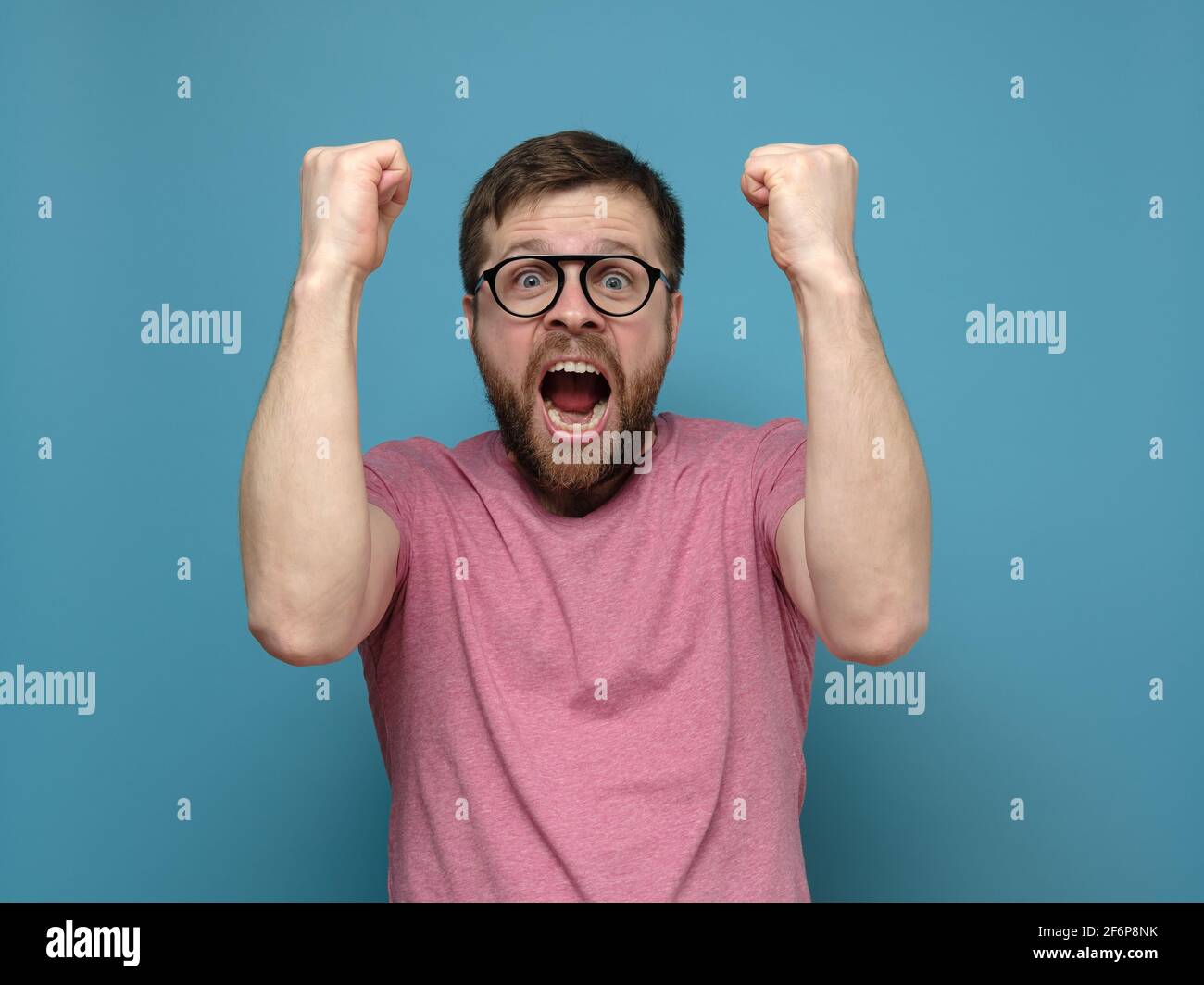 Ein emotionaler, glücklicher bärtiger Mann mit Brille und einem pinken T-Shirt macht eine Geste zum Sieger, schreit fröhlich und macht große Augen. Stockfoto