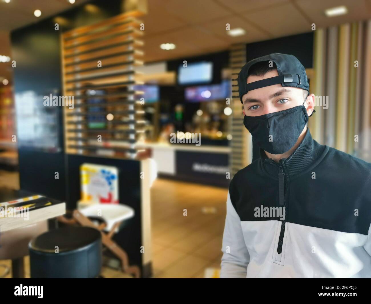 Junger Mann mit schwarzer Baseballmütze und Gesichtsmaske aufgrund von Covid19 und einer trendigen Sportjacke in einem Takeaway Restaurant mit seichtem Fokus. Stockfoto