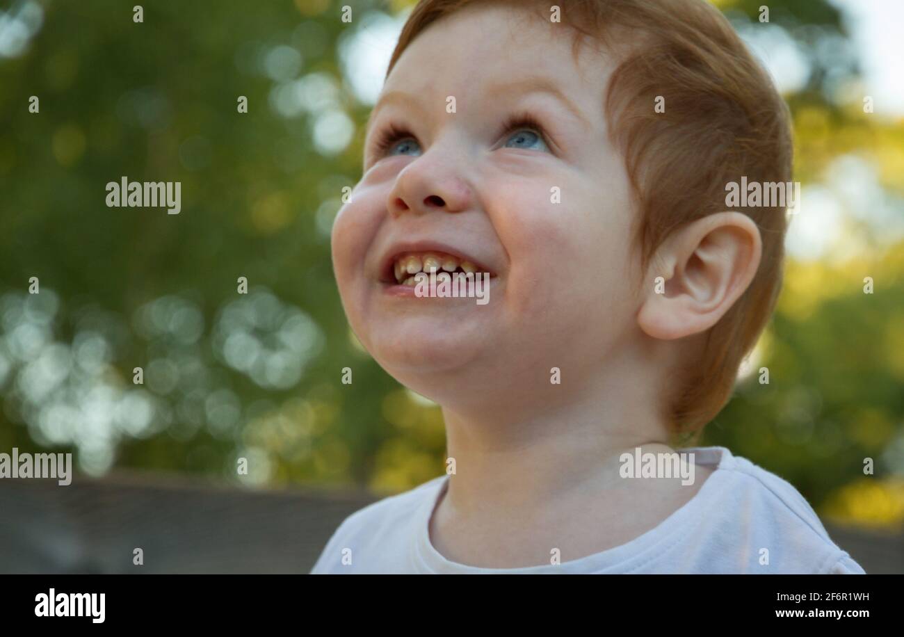 Porträt eines niedlichen, rothaarigen, blauäugigen Jungen in einem weißen T-Shirt auf einem Spielplatz an einem sonnigen Tag Stockfoto