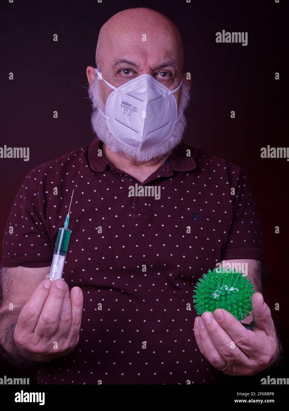 Leitender Mann mit Schutzmaske hält eine Impfstoffspritze in sich Eine Hand und eine grüne Kugel, die das Corona-Virus darstellt In einem anderen Stockfoto