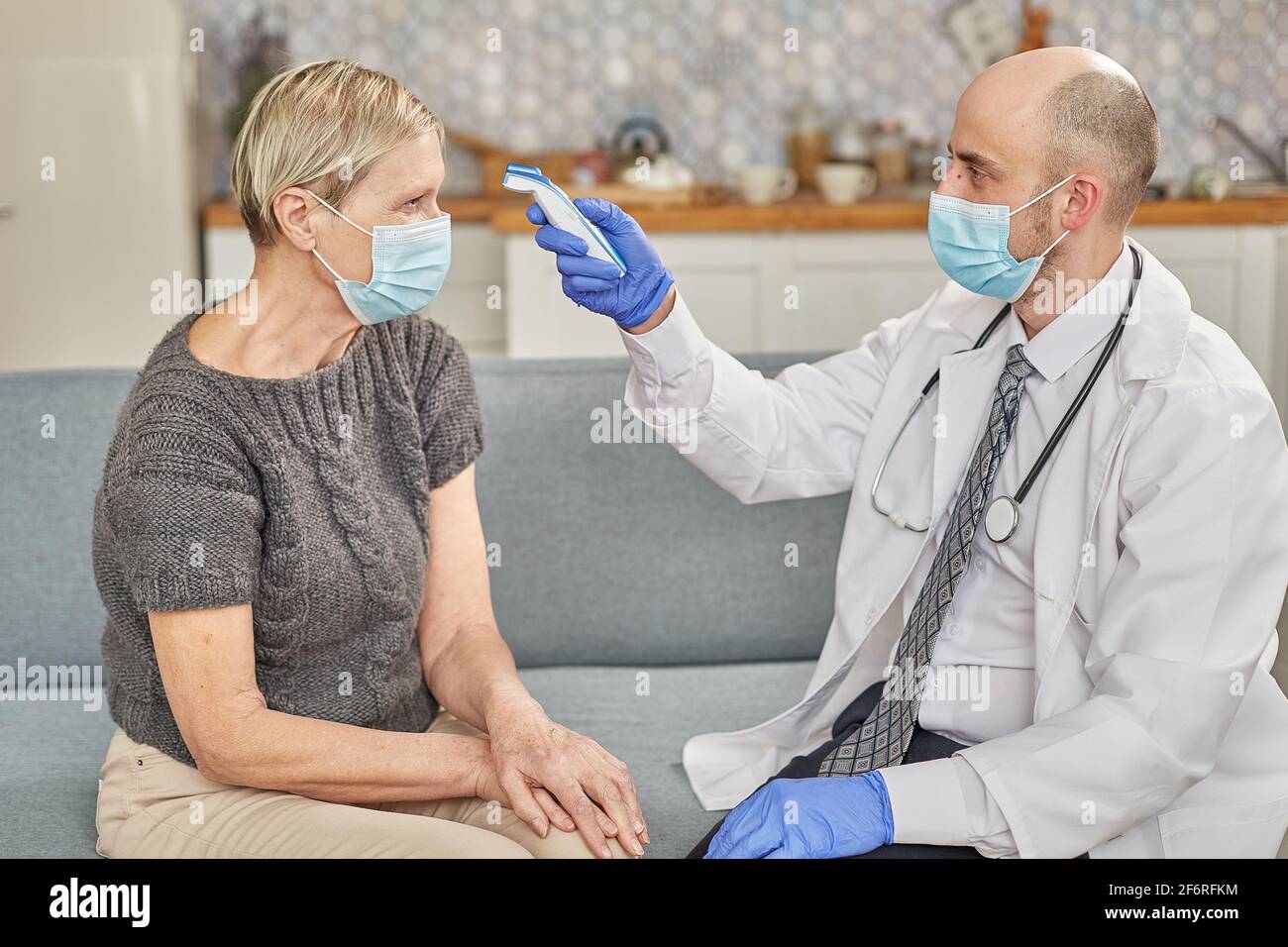 Ein Arzt misst die Temperatur einer älteren Frau in ihrem Zuhause mit einer Schutzmaske. Coronovirus-Infektionsdiagnosekonzept Stockfoto