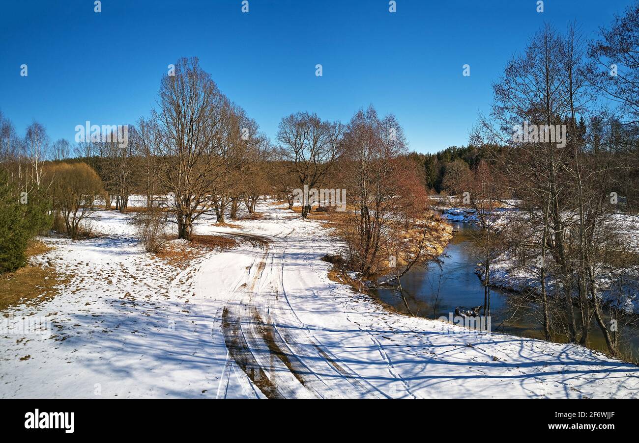 Frühlingshafte Landschaft mit schmelzendem Schnee und aufgetauten Flecken auf der Wiese. Wald und Isloch Bach im sonnigen märz april Tag. Schmutzige Landstraße, Eis und Schnee Stockfoto