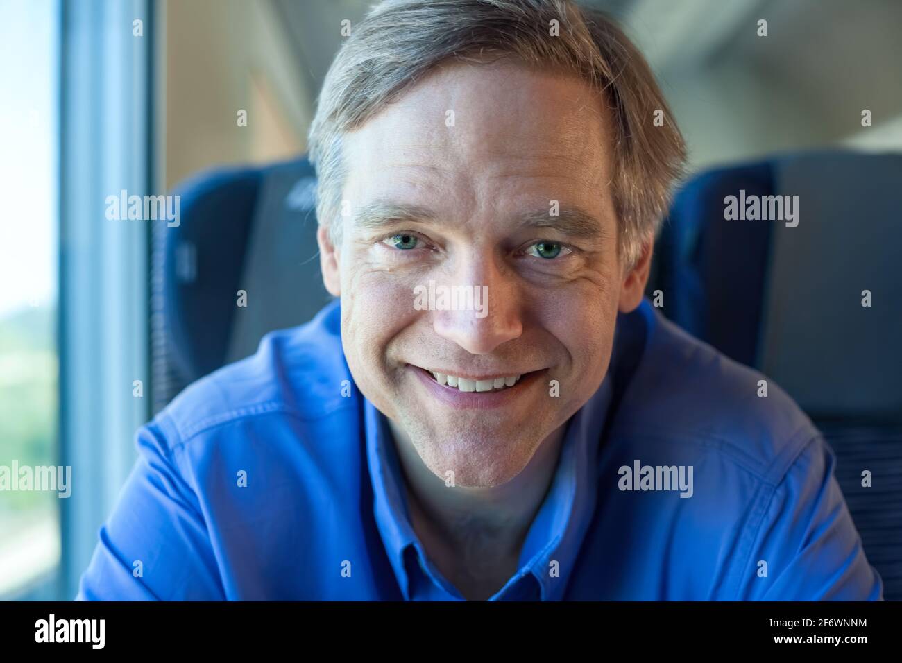 Kaukasischer Mann in den Vierzigern mit blauem Hemd im Pendlerzug, lächelnd und vor der Kamera sitzend Stockfoto