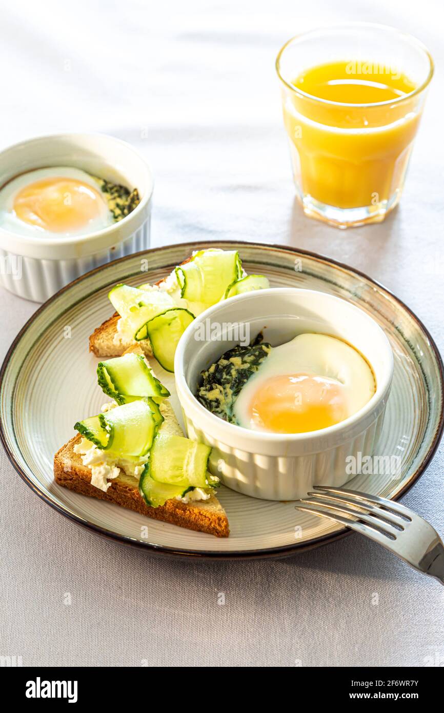 Geraffte Eier (Oeuf Cocotte) oder gebackene Eier. Gesundes Frühstück mit Eiern und Spinat und Toast mit Gurke Stockfoto