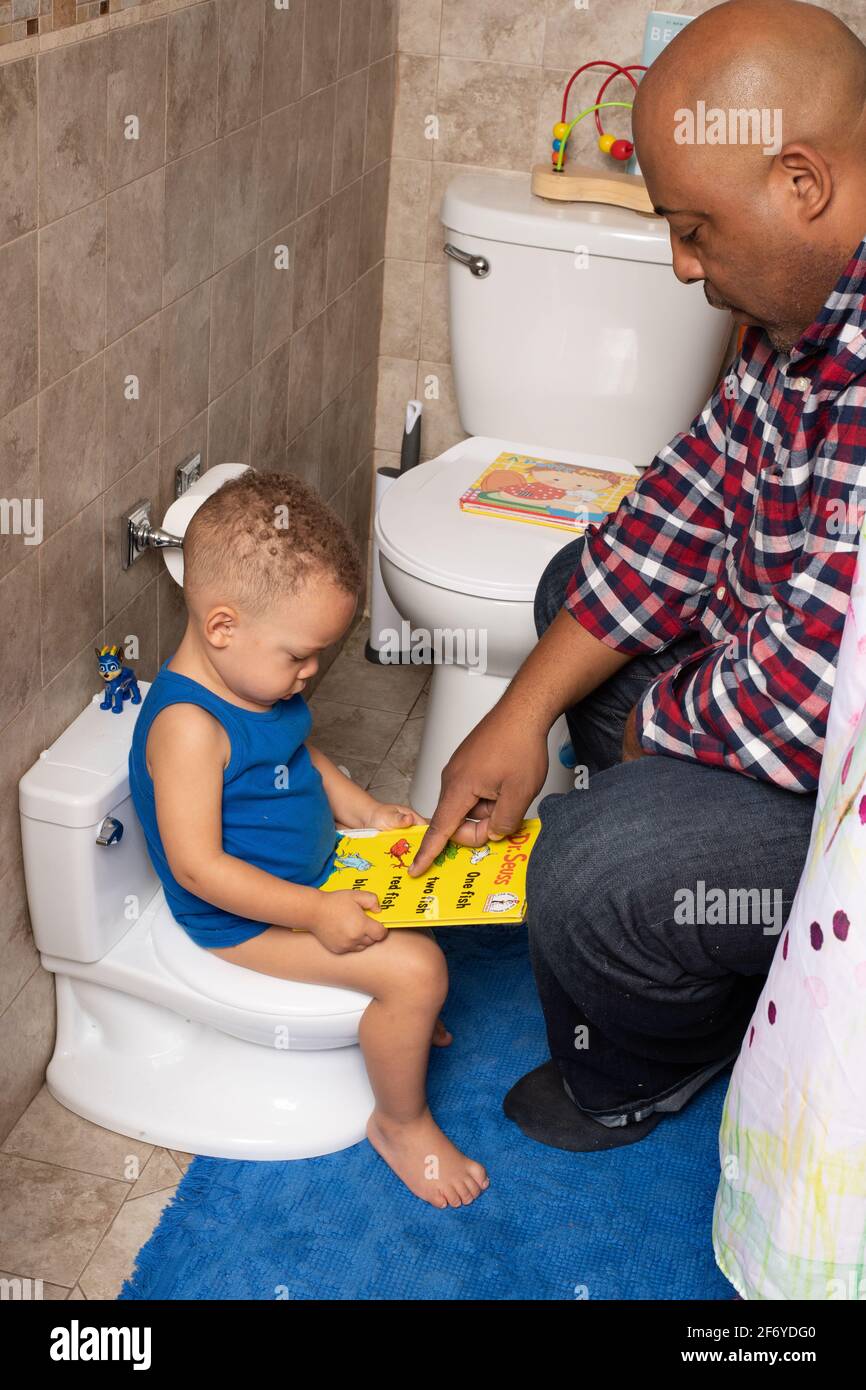 Toilettentraining: 2-jähriger Junge sitzt auf einem Töpfchen im Badezimmer und schaut sich mit dem Vater Bücher an Stockfoto