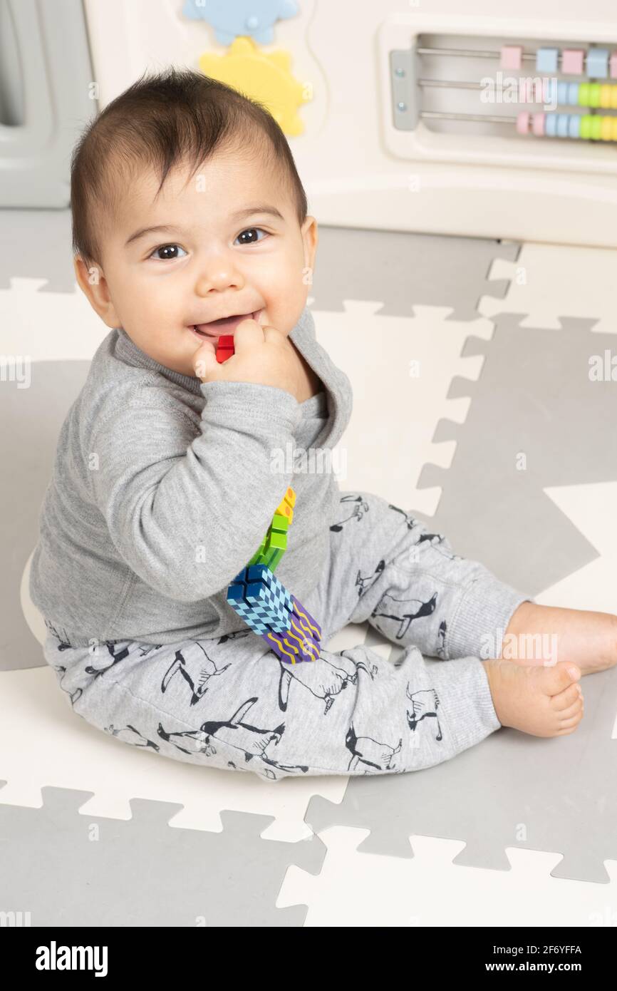 Sechs Monate alter Junge, der auf dem Boden sitzt und lächelt Blick auf die Kamera, die die Hand mit einem kleinen Spielzeug in der Nähe des Mundes hält Stockfoto