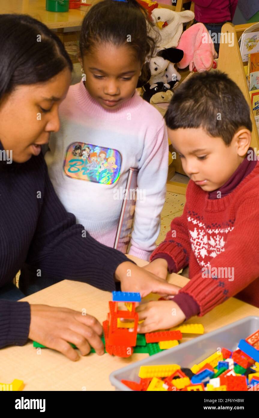 Vorschulklasse Klassenzimmer 3-4 Jahre alt Lehrerin im Gespräch mit Jungen Über die Struktur, die er mit Lego-Plastikziegeln gemacht hat Als Mädchen hält Buch aussieht Stockfoto