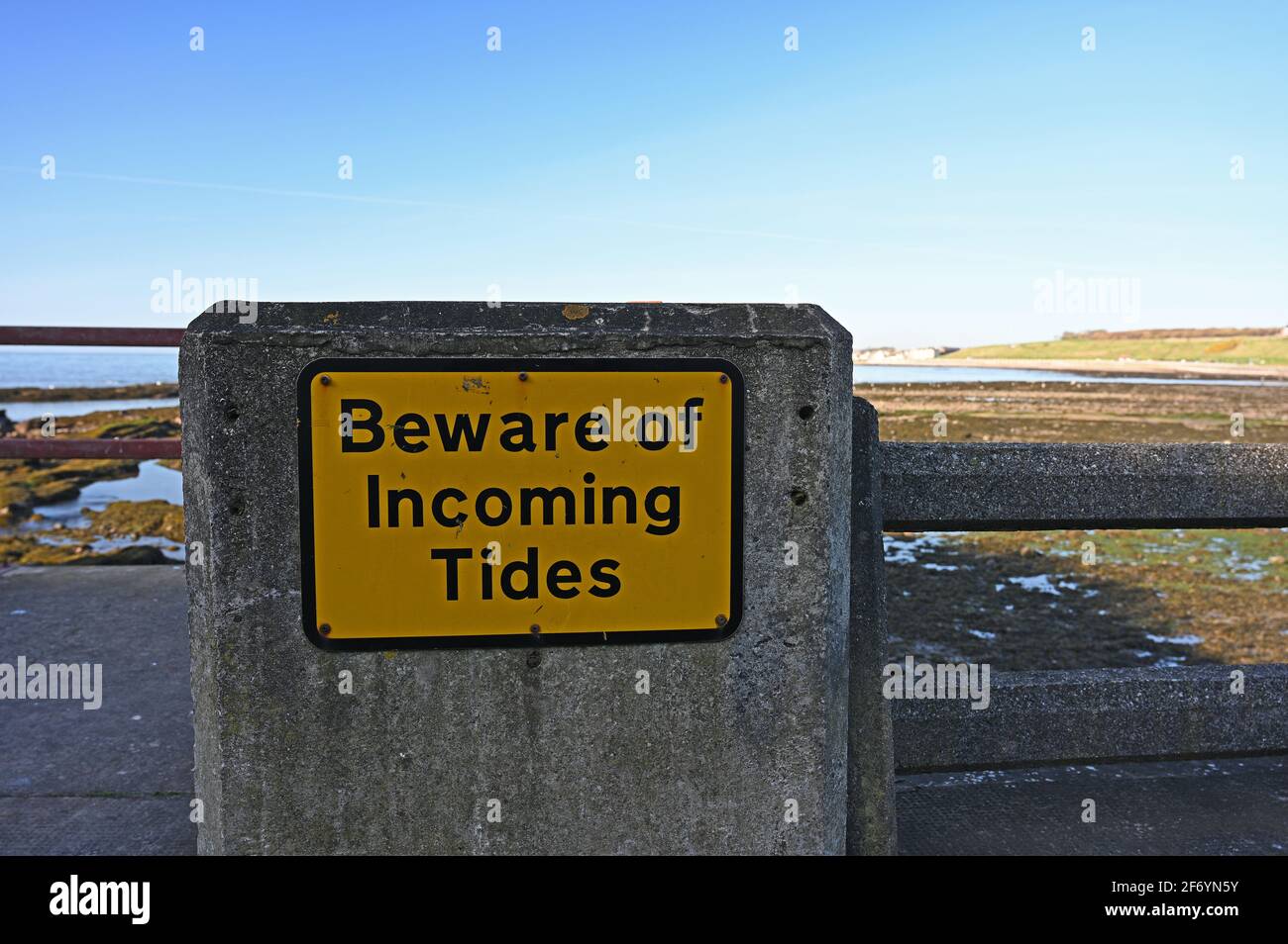 Verwittert Vorsicht vor dem eingehenden Tides-Zeichen, gelben und schwarzen rechteckigen Zeichen an der Küstenmauer mit verschwommenem Küstenhintergrund. Stockfoto