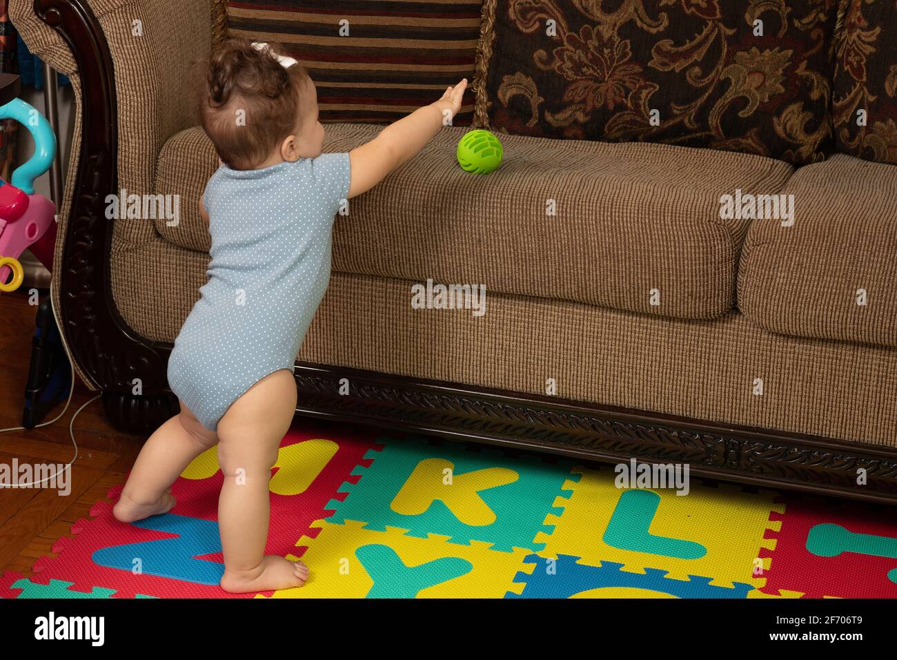 7 Monate altes Mädchen, das mit Unterstützung auf der Couch steht und nach dem Ball auf dem Kissen greift Stockfoto