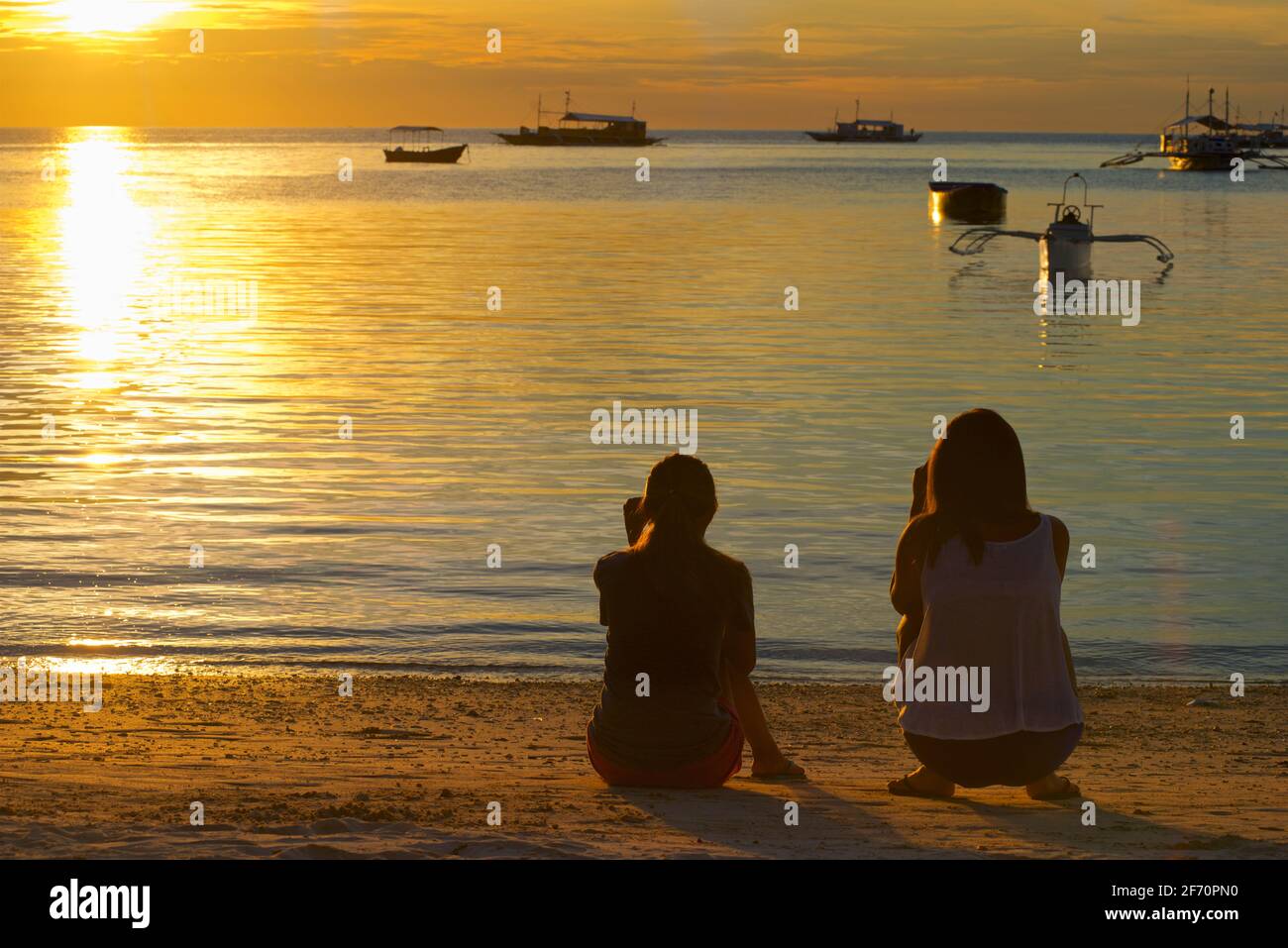 Silhouette zweier Freunde, die bei Sonnenuntergang am Strand am Ufer des Visayan-Meeres sitzen. Logon Village, Malapascua Island. Cebu, Philippinen Stockfoto