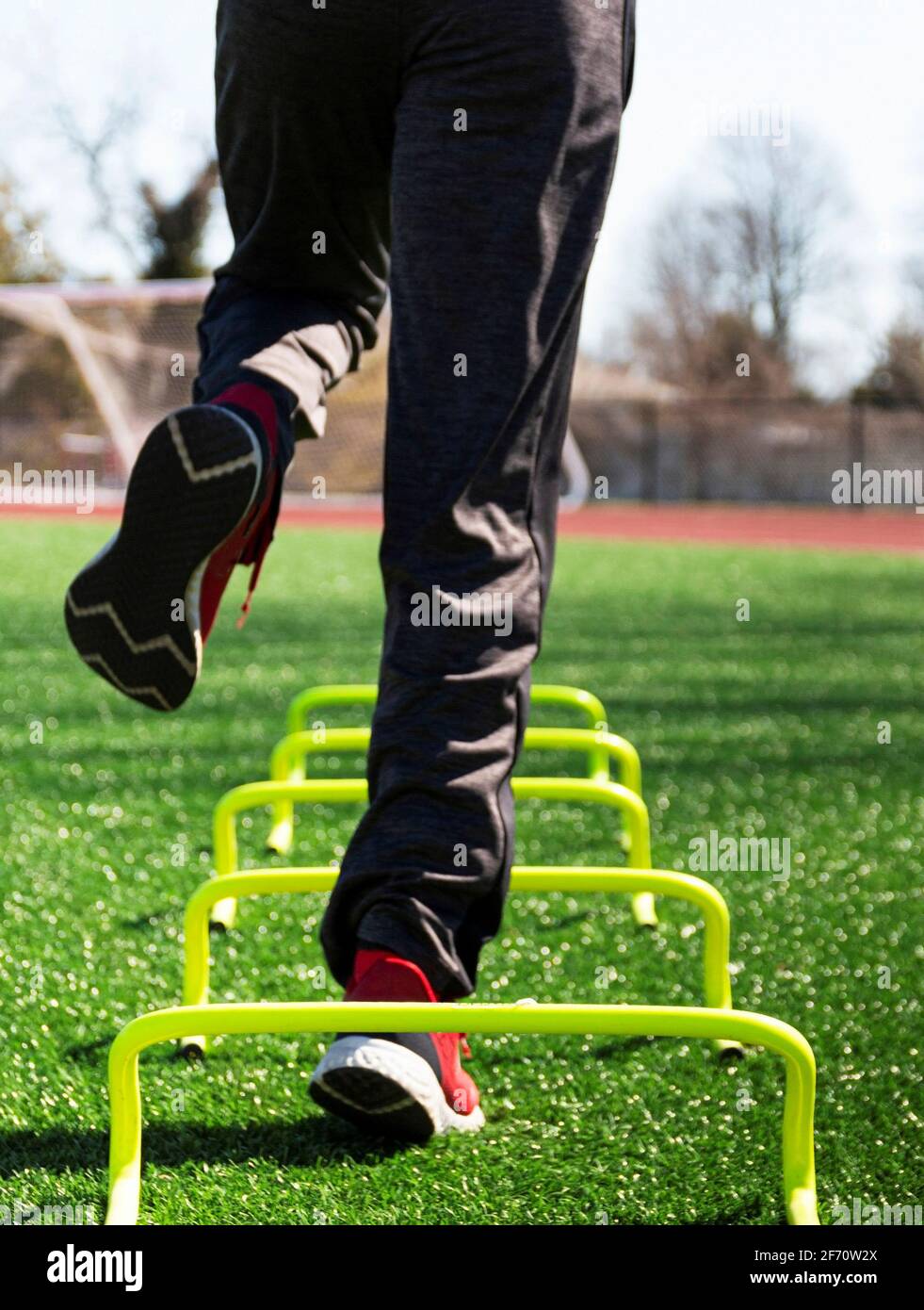 Low-Angle-Ansicht eines männlichen Athleten, der auf einem Bein über gelbe Mini-Hurldes auf einem grünen Rasenfeld von hinten springt. Stockfoto