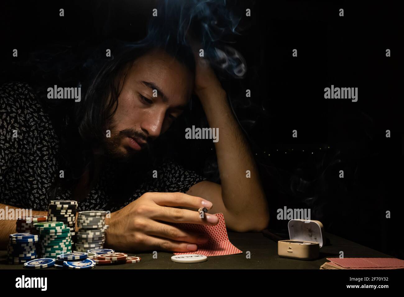 Mann, der Poker spielt, während er Karten betrachtet und Tabak raucht. Junger langhaariger Mann, Glücksspiel, dramatisches Licht. Stockfoto