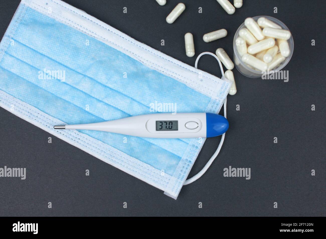 Weißes elektronisches Thermometer mit Packungen Tabletten und blauer medizinischer Gesichtsmaske auf schwarzem Hintergrund. Stockfoto