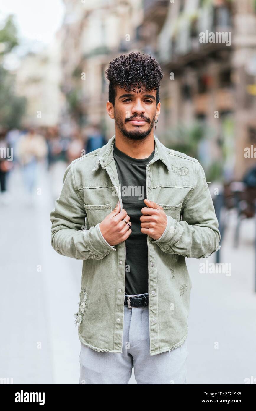 Vertikales Porträt eines jungen hispanischen Models, das in den Straßen von Barcelona posiert. Er kleidet sich in lässige grüne Kleidung und trägt Ohrringe und Ringe. Stockfoto
