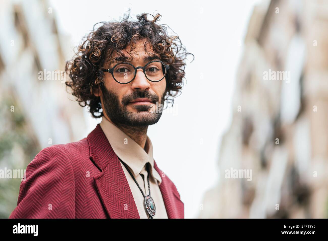 Horizontales Porträt eines jungen kaukasischen Mannes auf der Straße, der die Kamera anschaut. Tragen Sie trendige Freizeitkleidung und Brille. Er hat lockiges Haar und einen Bart Stockfoto