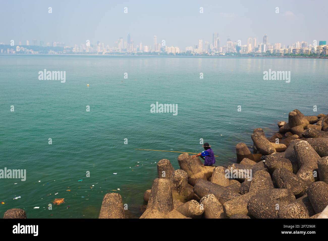 Ein Mann, der auf Tetrapoden-Stein am Meeresantrieb sitzt und mit Angelrute angeln kann. Müll schwimmt auf dem Wasser. Morgenansicht, Nariman Point, Mumbai. Stockfoto