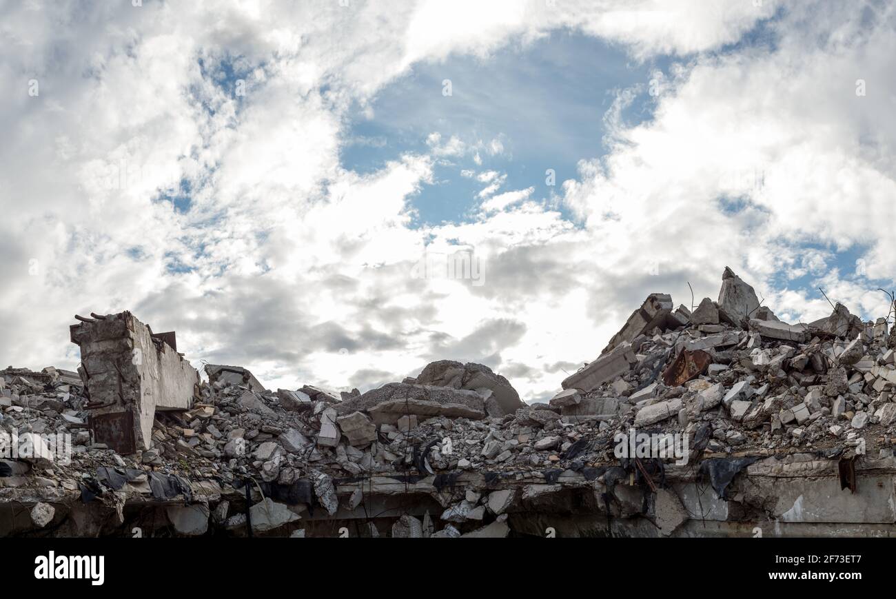 Ein Haufen aus grauen Betonschutt eines zerstörten Gebäudes mit einem riesigen Balken im Vordergrund gegen einen blauen Himmel mit Wolken. Panorama. Stockfoto