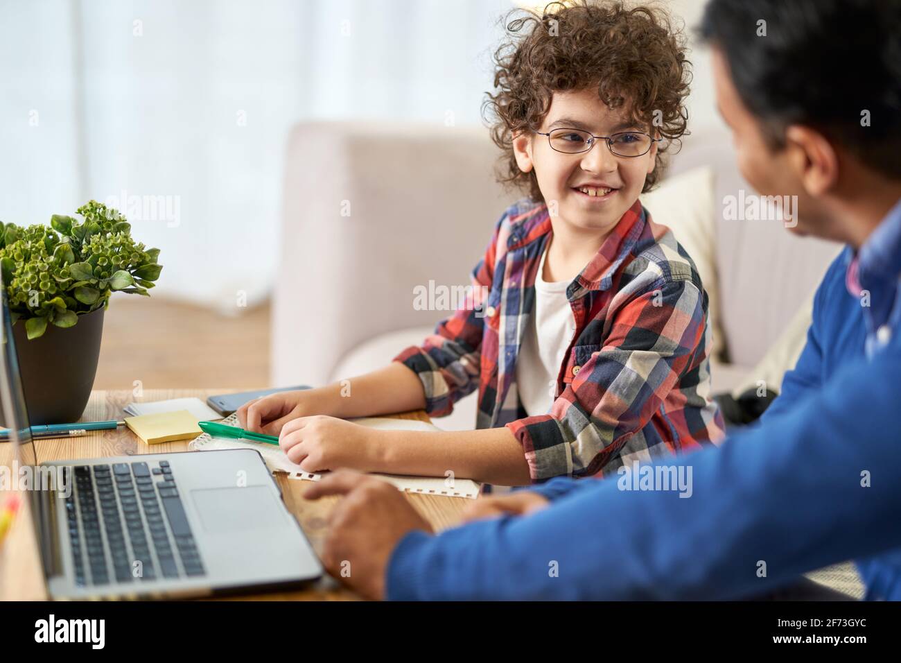 Sehr nette Schülerin. Fröhlicher kleiner lateinamerikanischer Schuljunge, der zusammen mit seinem Vater Hausaufgaben macht und dabei einen Laptop benutzt, während er zu Hause am Schreibtisch sitzt Stockfoto