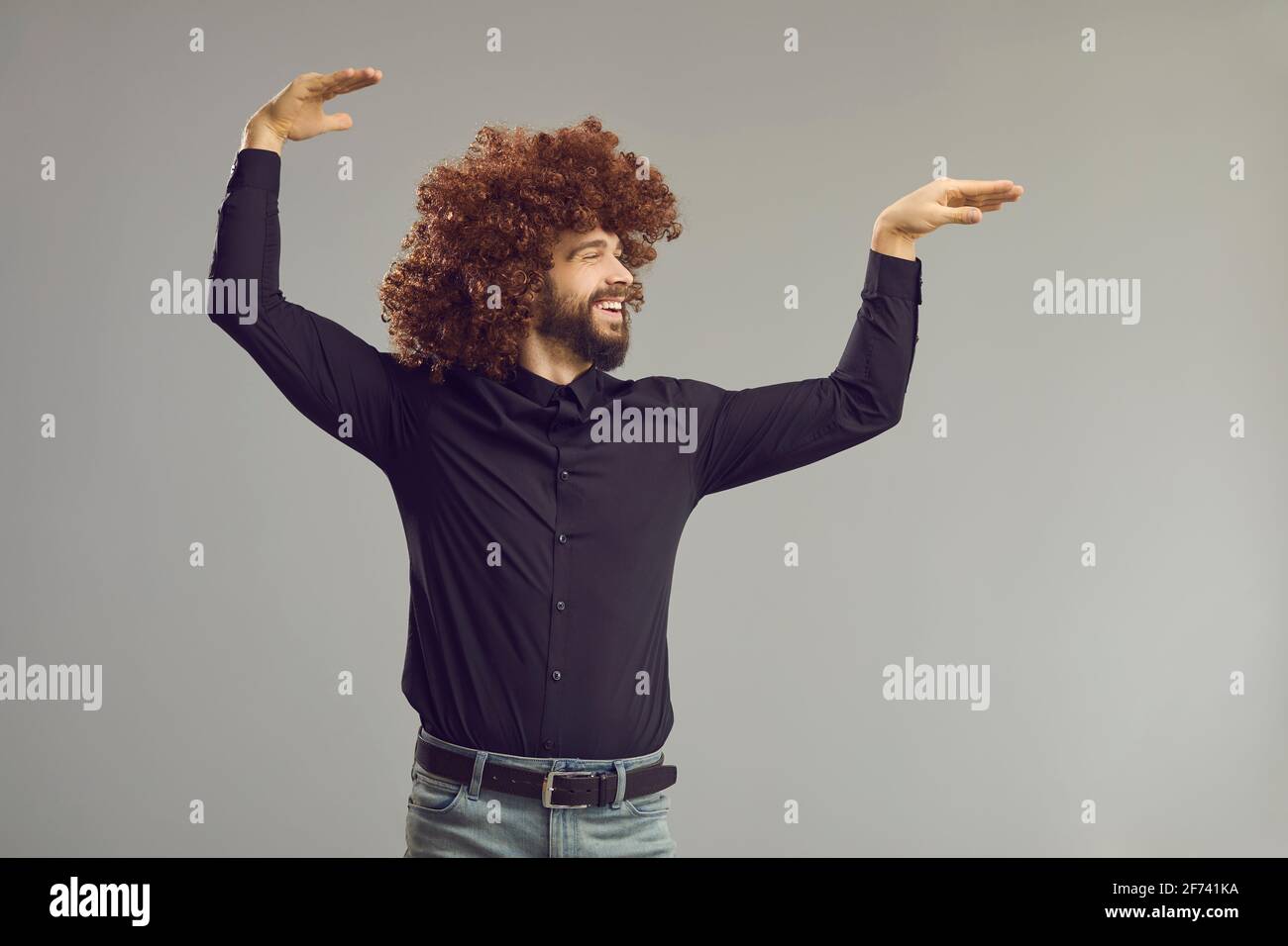Fröhlicher junger Mann mit verrückter, witziger lockiger Frisur, die tanzt Grauer Studiohintergrund Stockfoto
