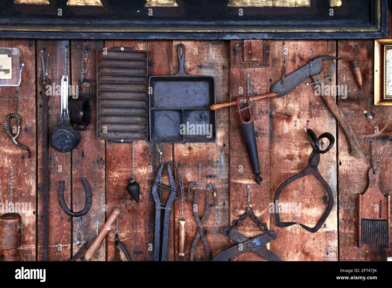 Rustikale Land alte antike Metall-Werkzeuge, Klemme, Rauhhhalsklaue, tong, Metallbecher, Hufeisen, hängende handgemachte Gegenstände, die an einer alten Scheunenwand aus Holz hängen Stockfoto