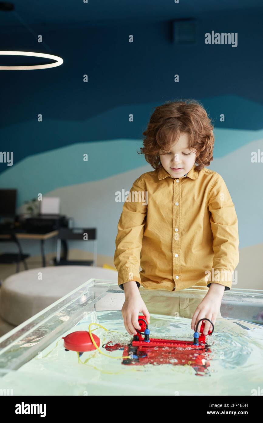 Vertikales Porträt von niedlichen lockig haarigen Jungen Putting Robo Boot Im Wasser beim Experimentieren mit Technologie im Robotik-Labor in Schule Stockfoto