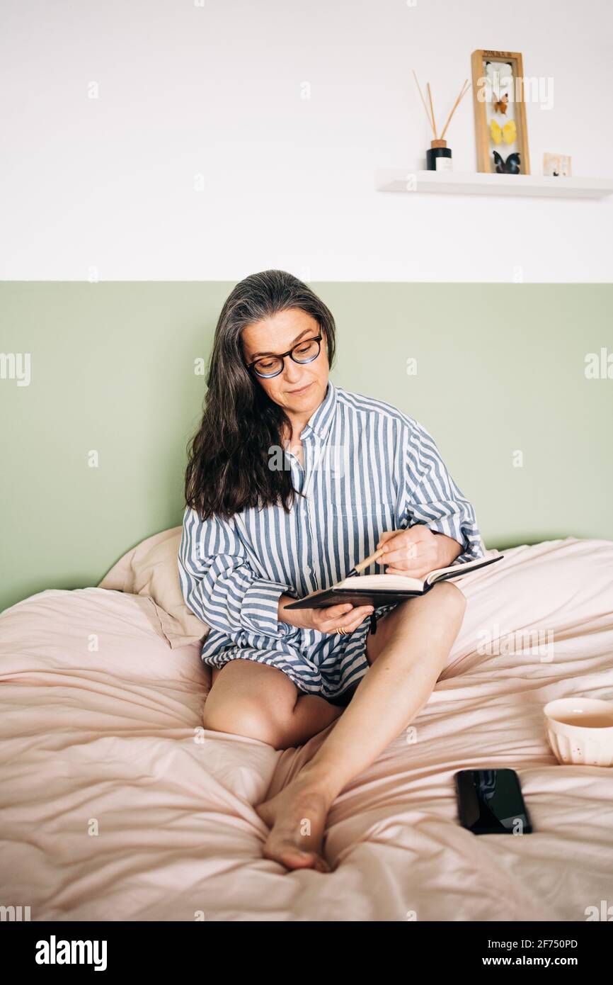 Fröhliche Frau mittleren Alters im gestreiften Hemd lächelnd und schreiend Notebook auf dem Bett mit Smartphone Stockfoto