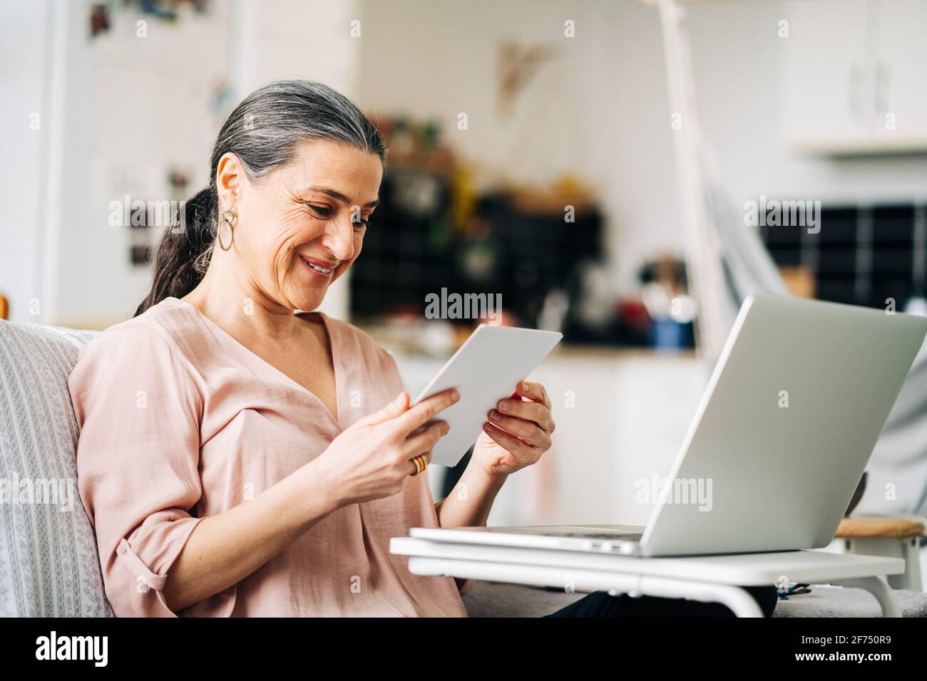 Fröhliche Frau mittleren Alters, die auf der Couch in der Nähe eines modernen Netbooks sitzt Beim Blick auf Foto in einer modernen Wohnung mit Küche an Unscharfer Hintergrund Stockfoto