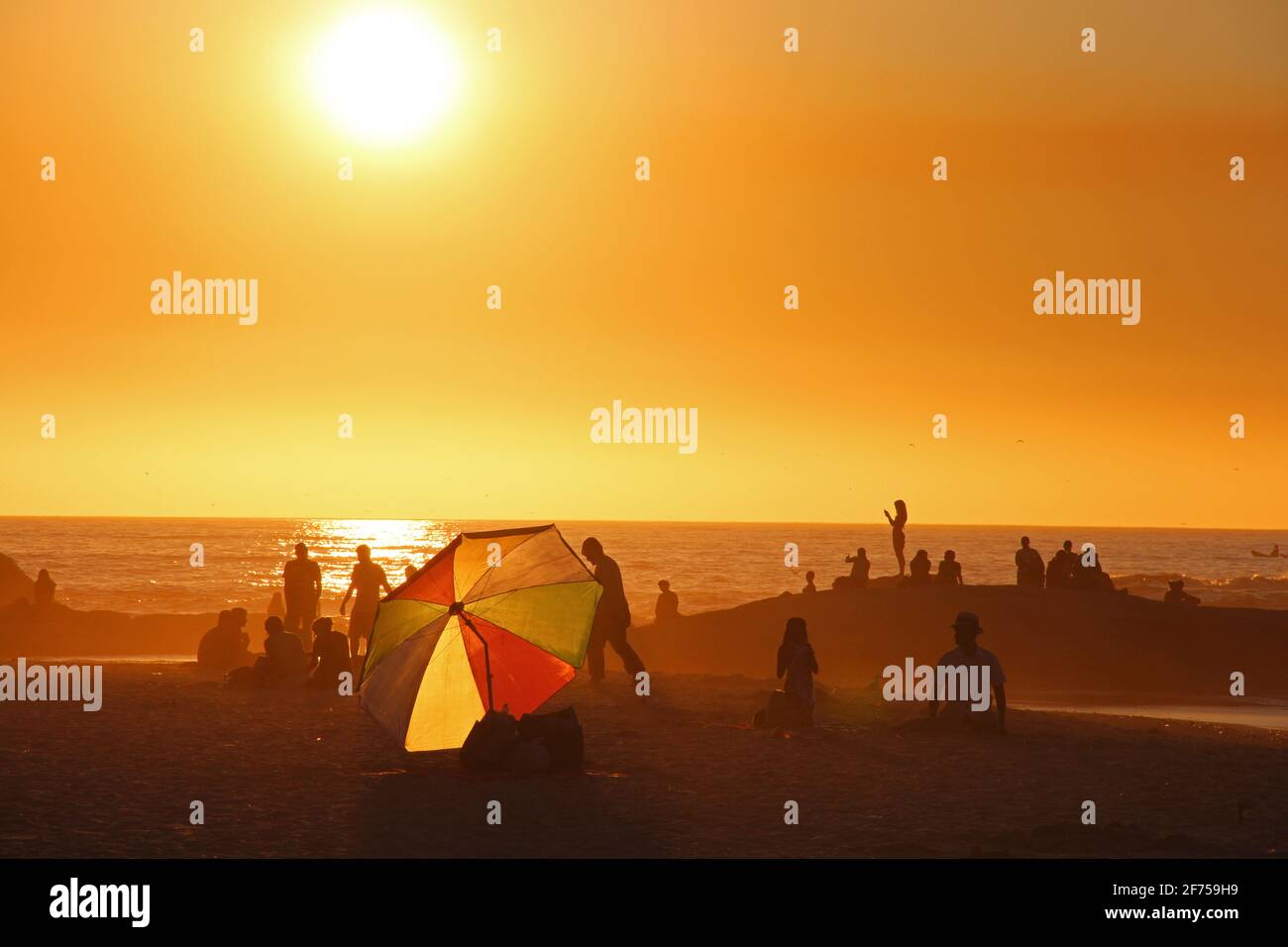 Lebhafte Sommerstrandszene in Kapstadt, Südafrika während der goldenen Stunde mit Silhouette von Menschen und buntem Sonnenschirm vor Sonnenuntergang Stockfoto