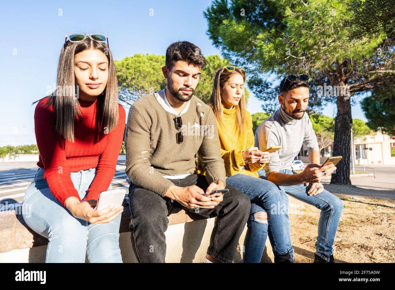 Eine Gruppe ernsthafter junger Menschen, die auf einer Stadtmauer sitzen und Zeit mit dem Smartphone mit mobiler Verbindung verbringen. Neue normale menschliche Gewohnheiten und gegenseitige Relatio Stockfoto