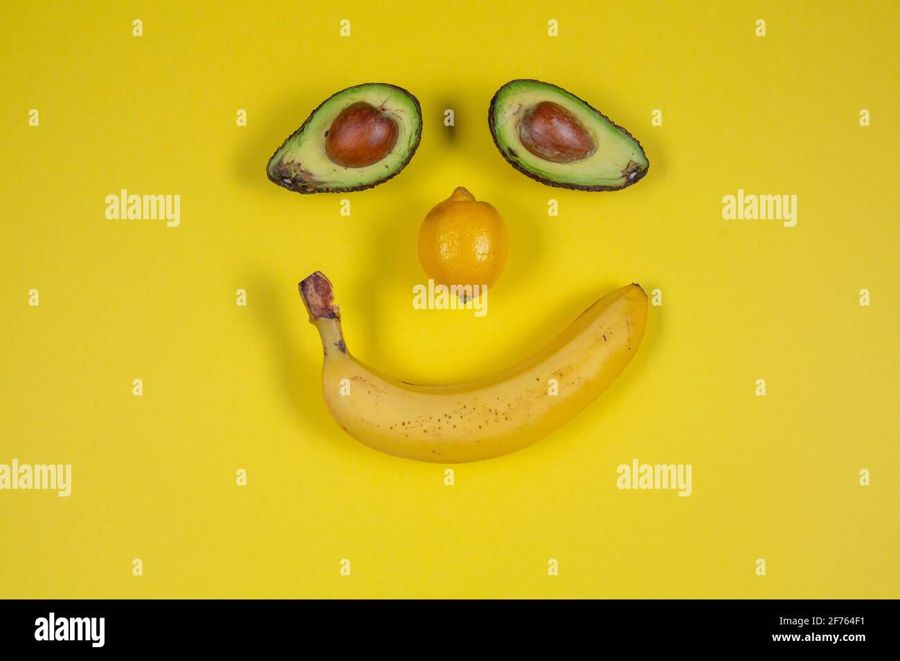 SCHÜCHTERNES GESICHT AUS BANANE, AVOCADO UND ZITRONE AUF GELBEM HINTERGRUND Stockfoto