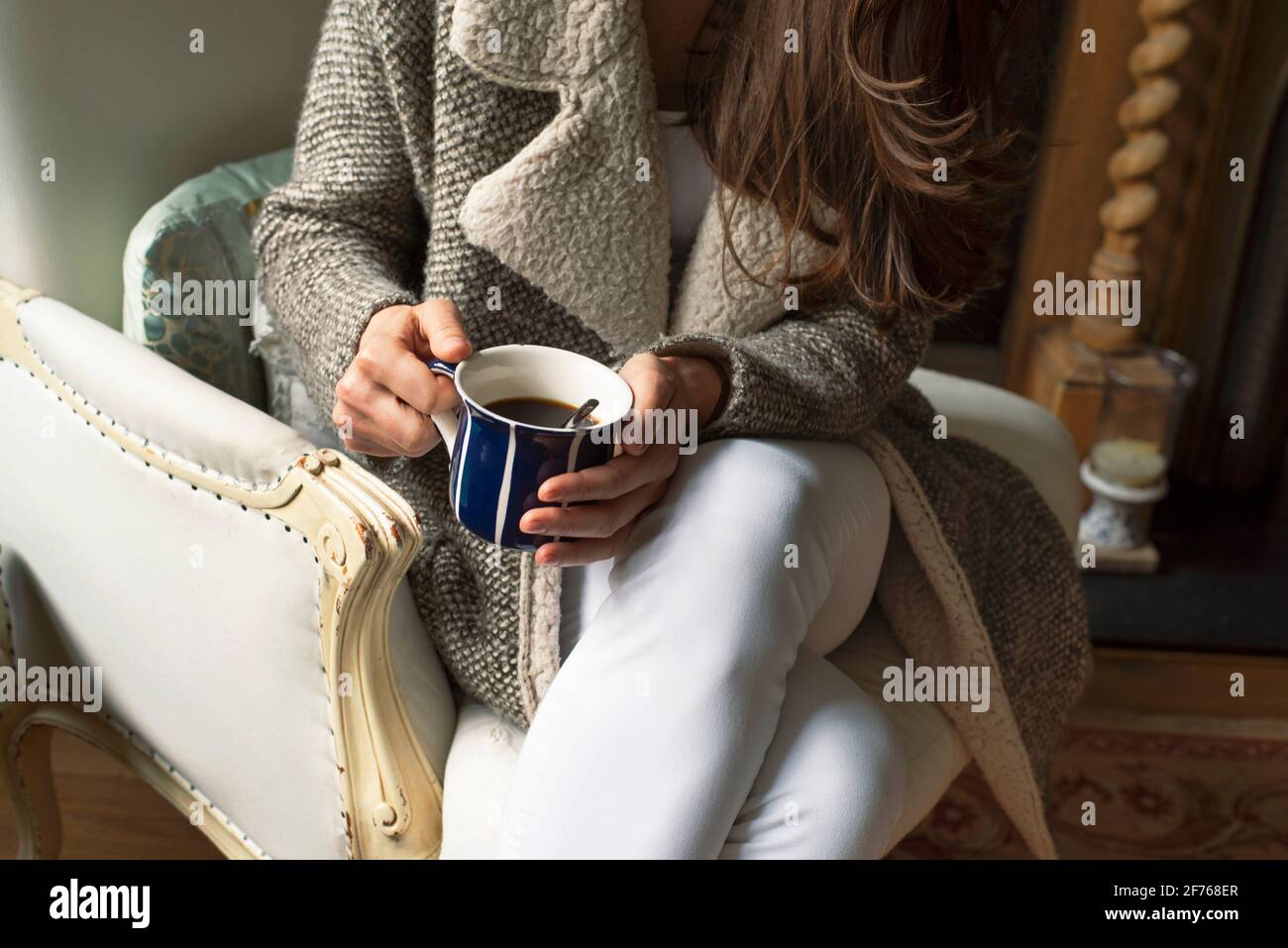 Nahaufnahme einer Frau in warmer Strickmode, die mit einer Tasse Kaffee auf einem Ledersessel sitzt. Indoor Freizeit Lifestyle mit rustikalem Landhausstil. London, Großbritannien Stockfoto