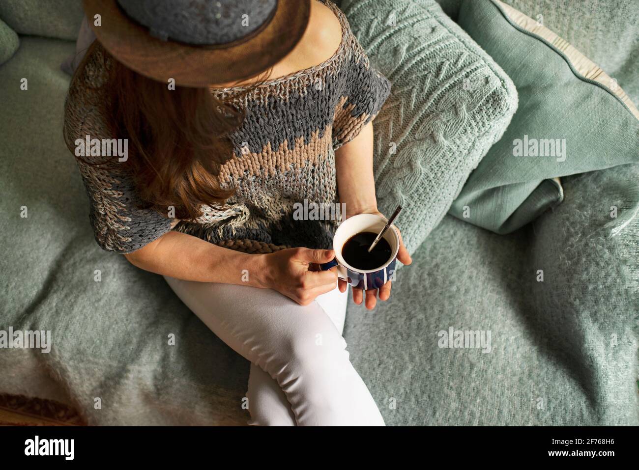 Draufsicht auf Frau in stilvoller Strickmode, die auf einer gemütlichen Enteneiercouch sitzt und eine Tasse Kaffee hält. Indoor Lounge auf Sofa Lifestyle, Kaffeepause Konzept. Stockfoto