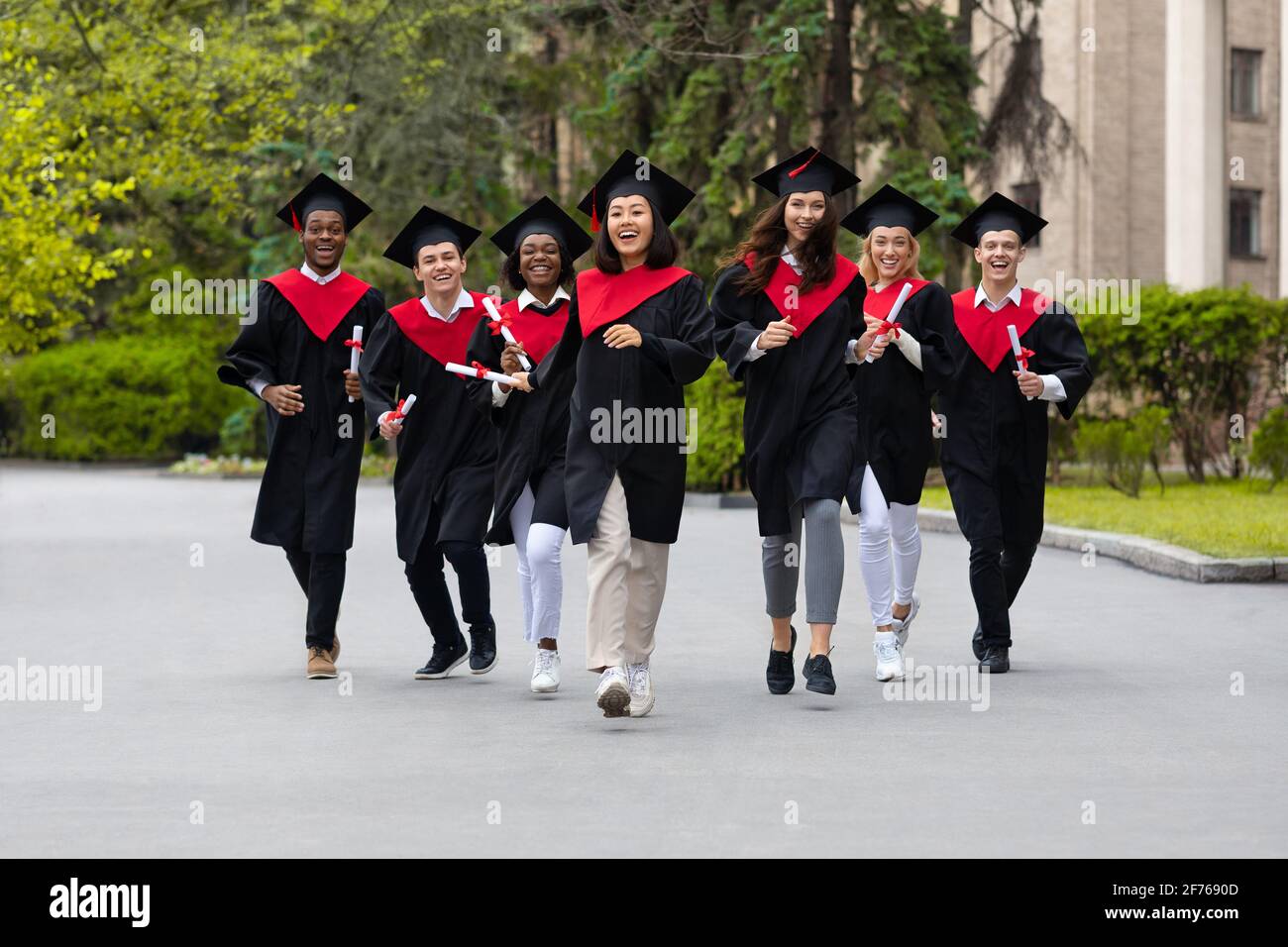 Fröhliche, multirassische Studenten in Abschlusskostümen, die durch den Universitätscampus laufen Stockfoto