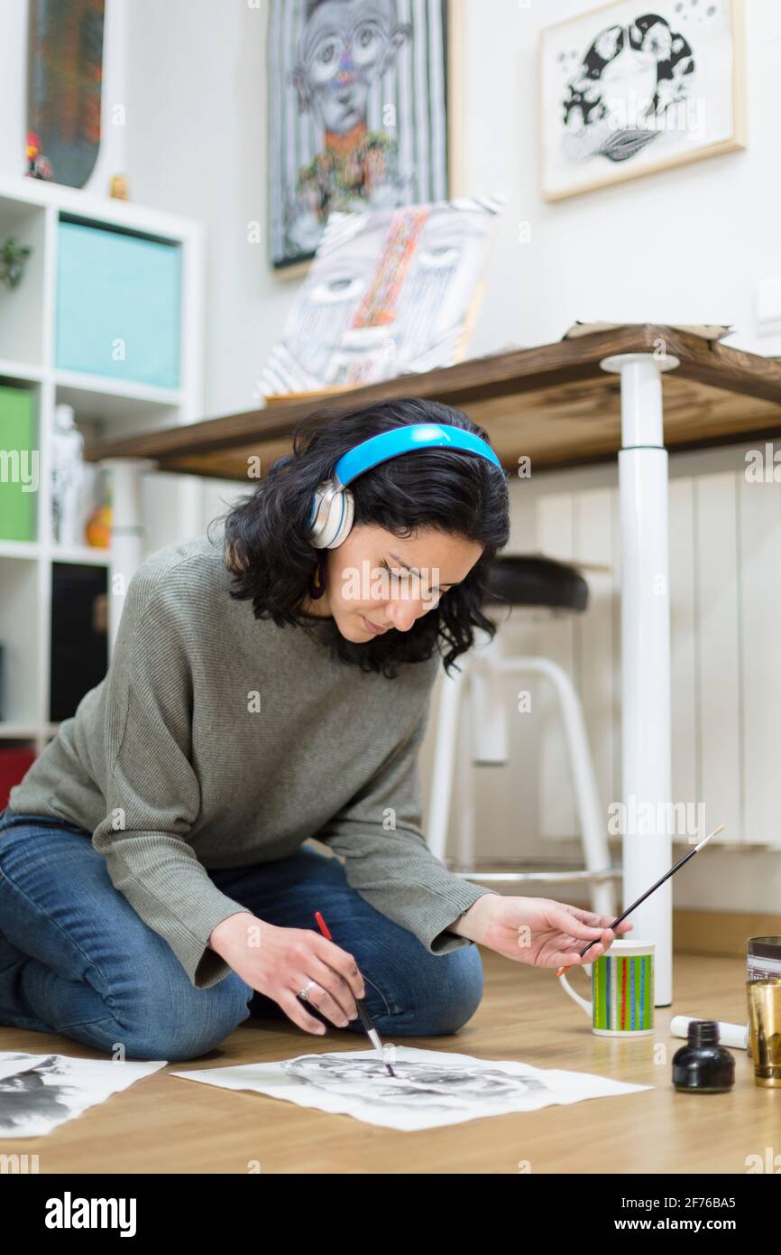 Künstlerin sitzt auf dem Boden und malt mit einem Pinsel auf Papier. Sie wird unterhalten und hört Musik. Stockfoto