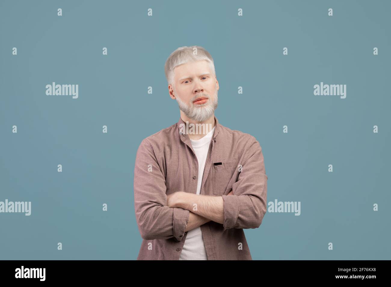 Junger Albino-Mann posiert mit gekreuzten Armen und schaut mit Ernsthafter Gesichtsausdruck bei der Kamera auf türkisfarbenem Hintergrund Stockfoto