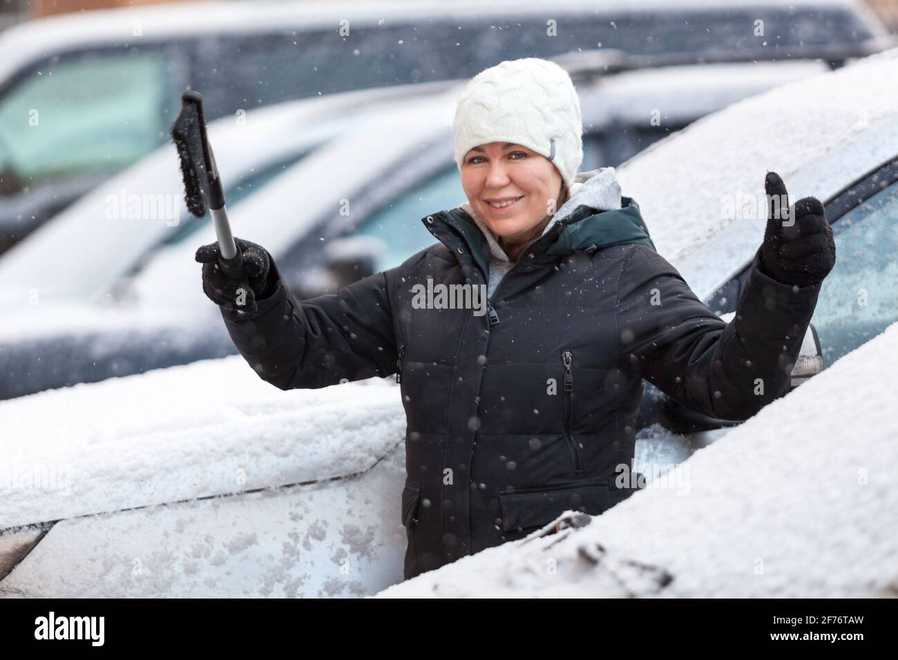 Frau bereit, ihren verschneiten Wagen nach Schneesturm zu reinigen, lächelnd und zeigt den Daumen nach oben Stockfoto